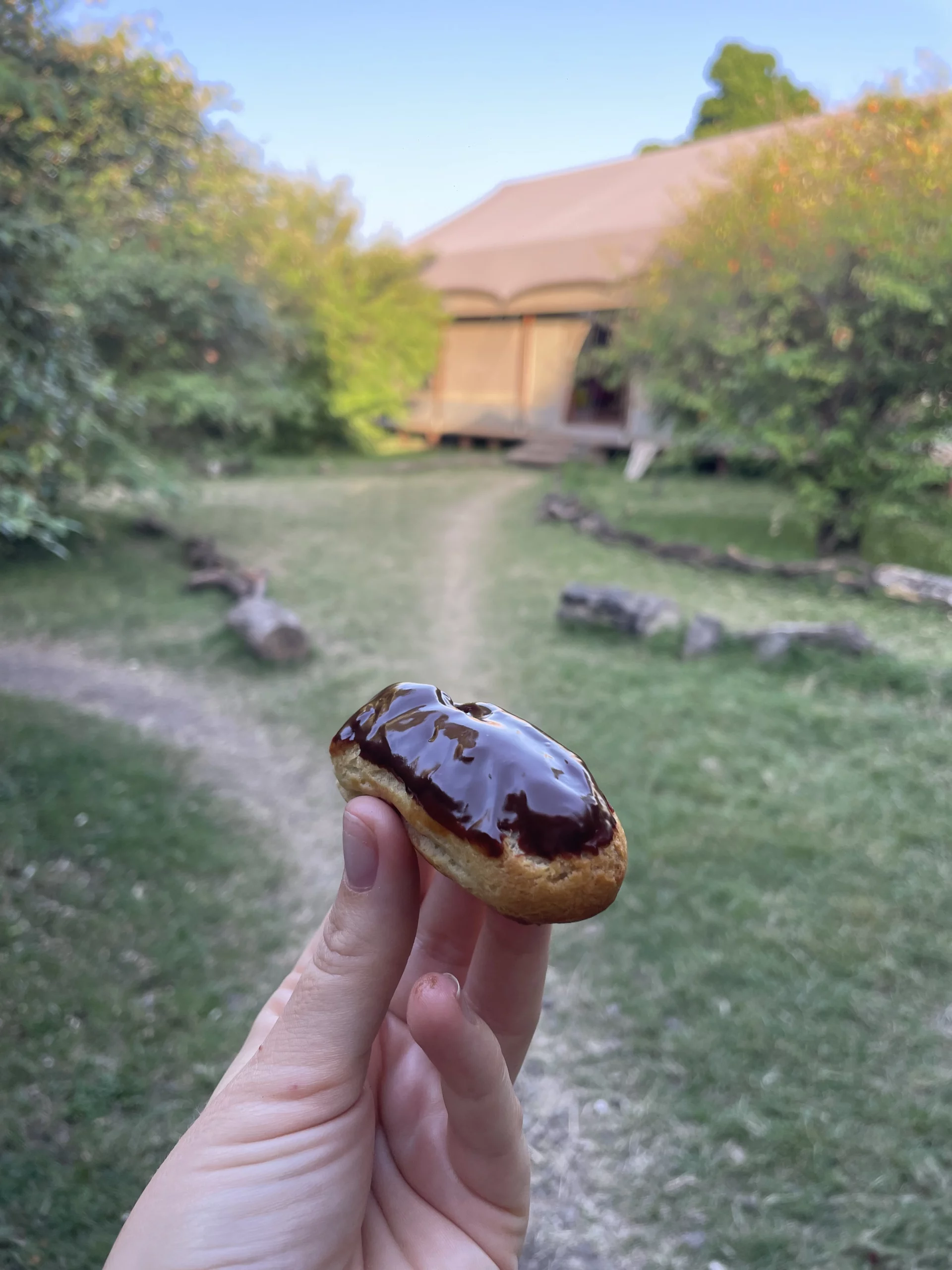 enkusero-kenya-eclair-tented-camp Chocolate-glazed éclair held in front of a tented camp — service-ready finish
